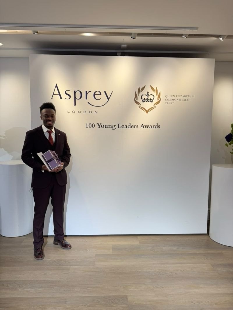 Elly Savatia, in a burgundy suit and red tie, smiles and holds a wrapped gift box in front of a white wall reading Asprey London, Queen Elizabeth II Commonwealth Trust, 100 Young Leaders Awards.