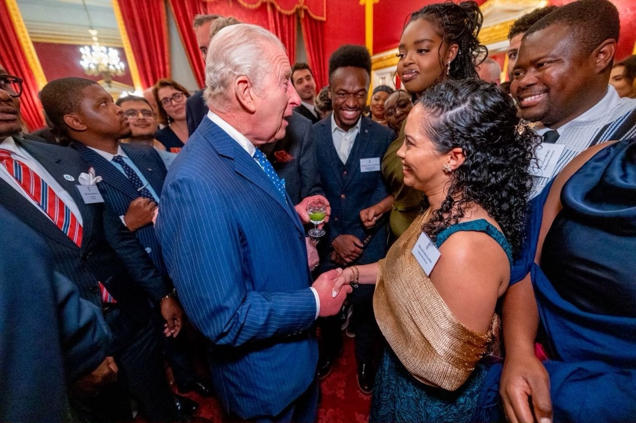A crowded reception in an ornate red and gold room. Young leaders wearing name badges smile as an older man in a pinstripe suit shakes hands with one of them.