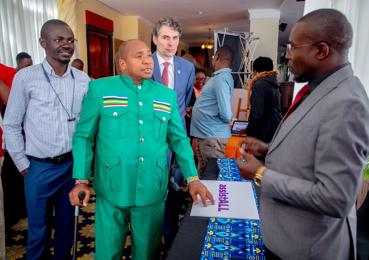 Hon. David Ole Sankok, in a green suit with East African flag pocket details and holding a cane, stands at an exhibition table with an assistALL sign. Three men stand around him in a busy hotel hall.