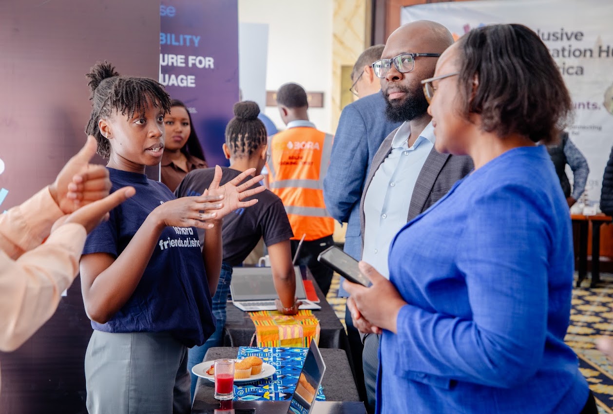 A young woman in a navy friends of ishara t-shirt explains her work with open hands to two delegates at an exhibition table with laptops and refreshments.