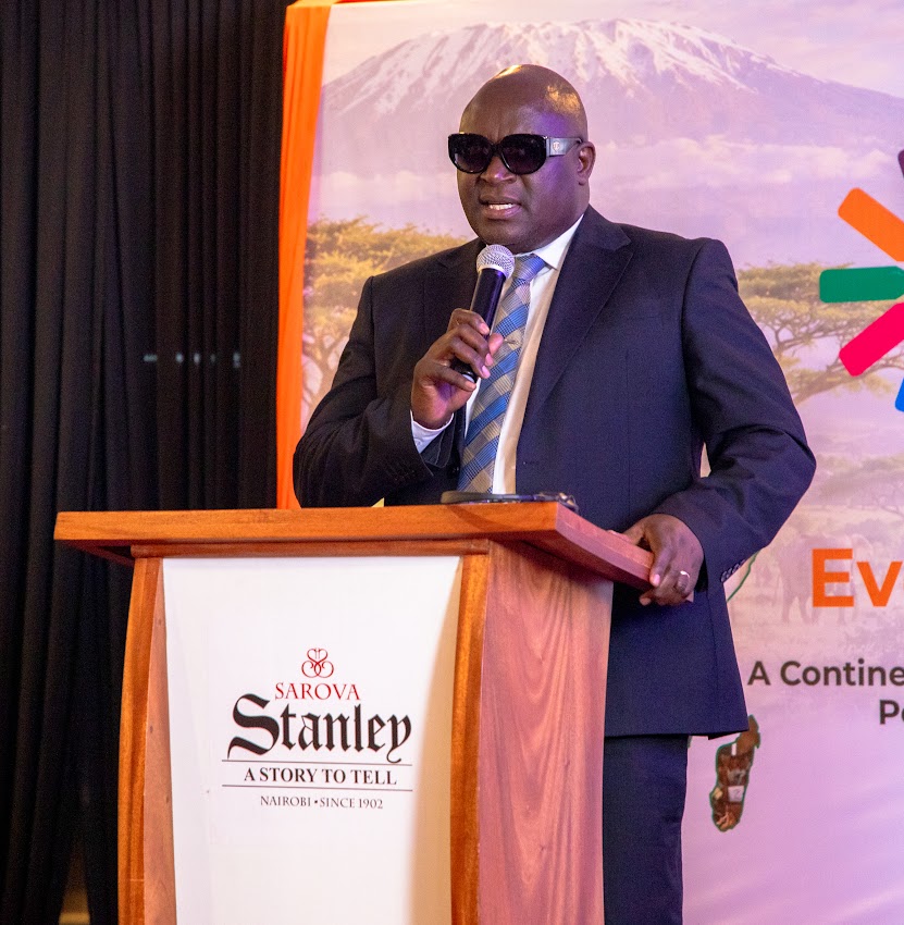 Mr. Fredrick Haga, in a dark suit and dark glasses, speaks into a microphone at a Sarova Stanley lectern.