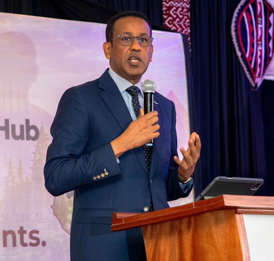 The Ambassador of Somalia to Kenya, in a navy suit and glasses, gestures as he speaks into a microphone in front of a purple banner.