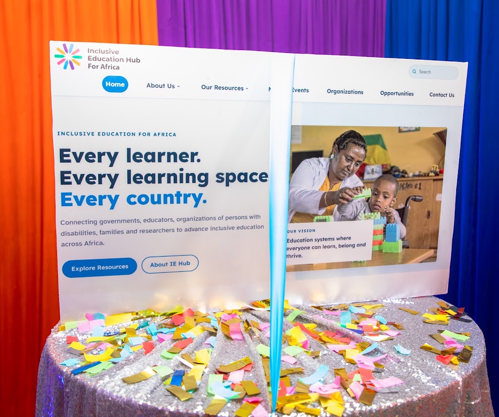A large board showing the Inclusive Education Hub for Africa homepage, with the headline 'Every learner. Every learning space. Every country.', on a table scattered with confetti in front of orange, purple and blue drapes.