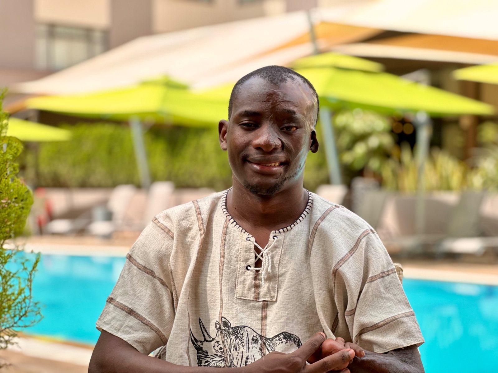 Haya Denis, in a cream linen shirt with a rhinoceros print, smiles at the camera beside a hotel swimming pool with green umbrellas behind him.