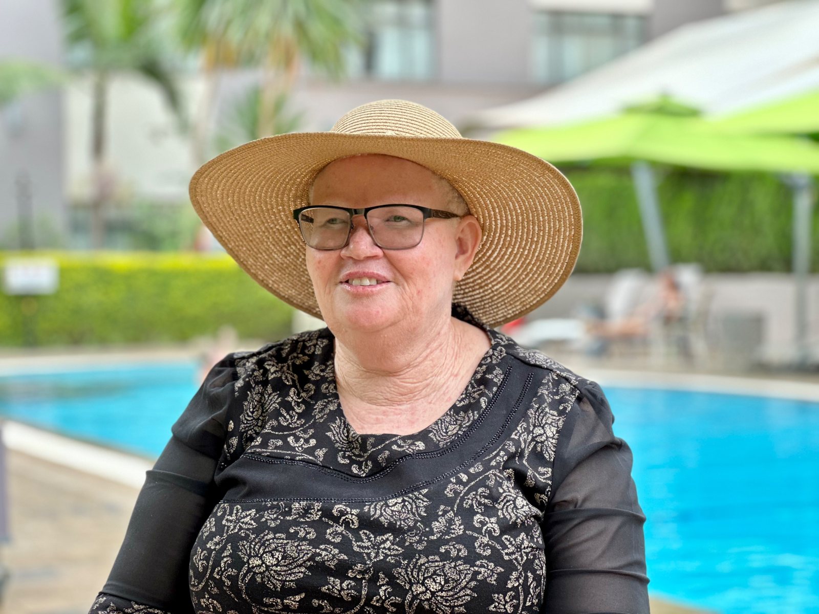 Jeriter Mutinda, wearing a wide straw hat, glasses and a black and gold patterned top, smiles beside a hotel swimming pool.