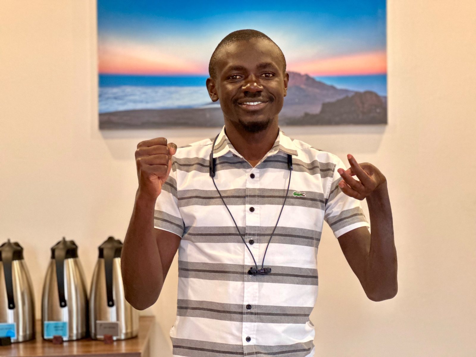 Joseph Owino, in a white and grey striped shirt, smiles and raises both hands in front of a painting of a mountain at sunrise.